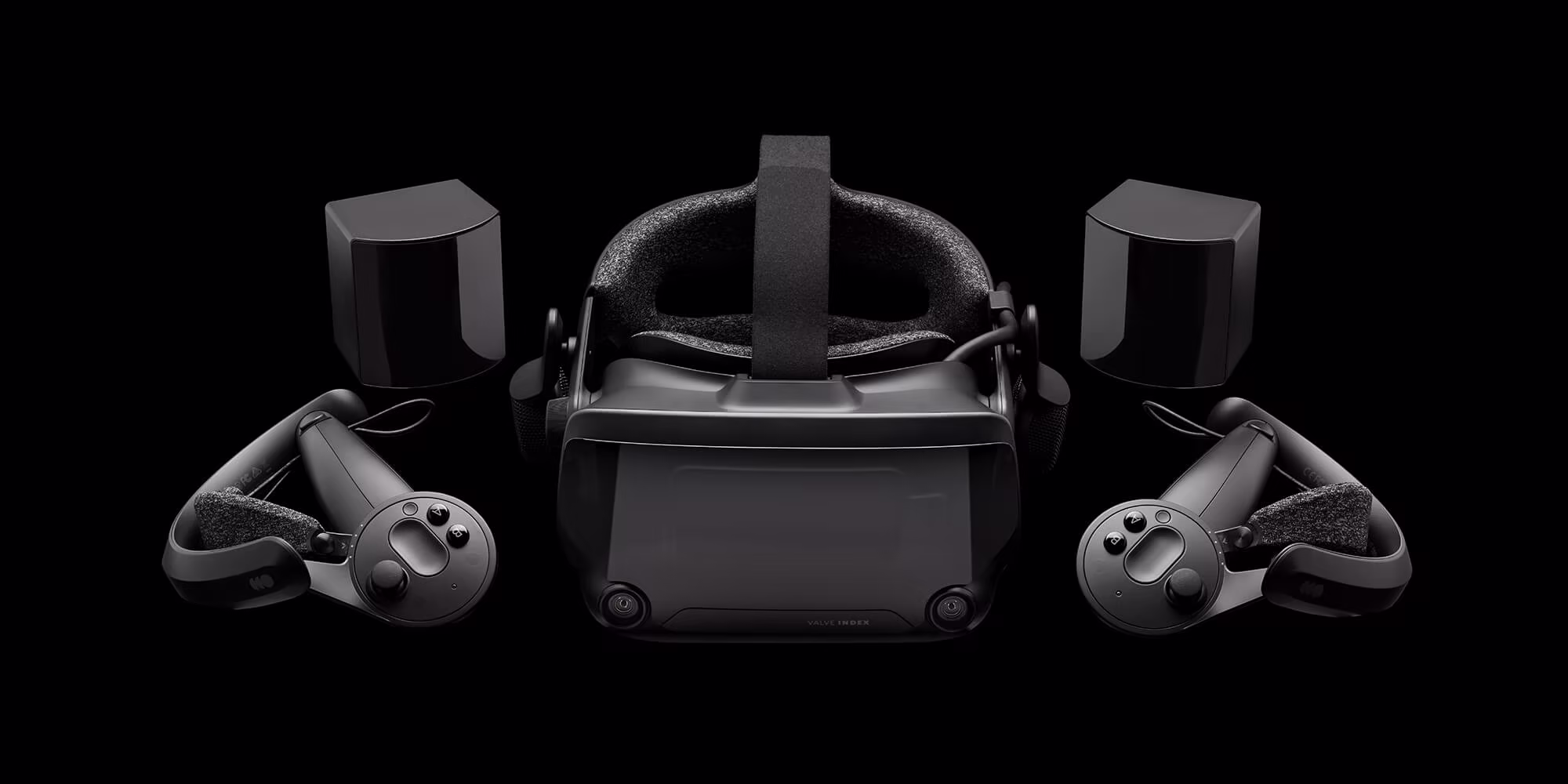 A Valve Index headset photographed head on against black, flanked by two Index controllers with their knuckle straps open and two SteamVR base stations